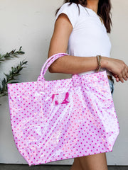 A person carrying a large Lulu Mac brand tote bag with a high-gloss finish. The bag features a light pink background covered in a pattern of small magenta polka dots, matching pink structured top handles, and a rectangular magenta patch in the center with the words "LULU MAC" printed in white text. The person holding the bag is wearing a white t-shirt and gold beaded bracelets.