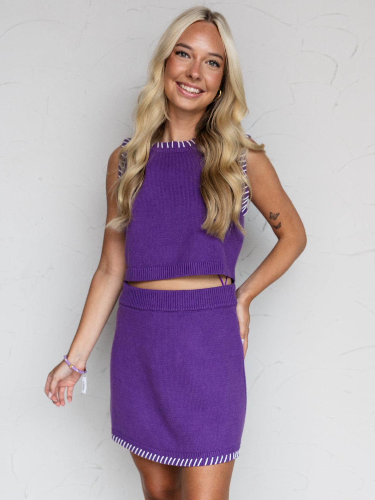 A full-length photograph of a smiling blonde woman posing with one hand on her hip against a textured white wall. She is wearing a matching two-piece purple knit set made from a 40% acrylic and 60% cotton fabric blend, featuring a sleeveless crop top and a mini skirt accented with white blanket-stitch details along the borders, styled with white western cowboy boots.