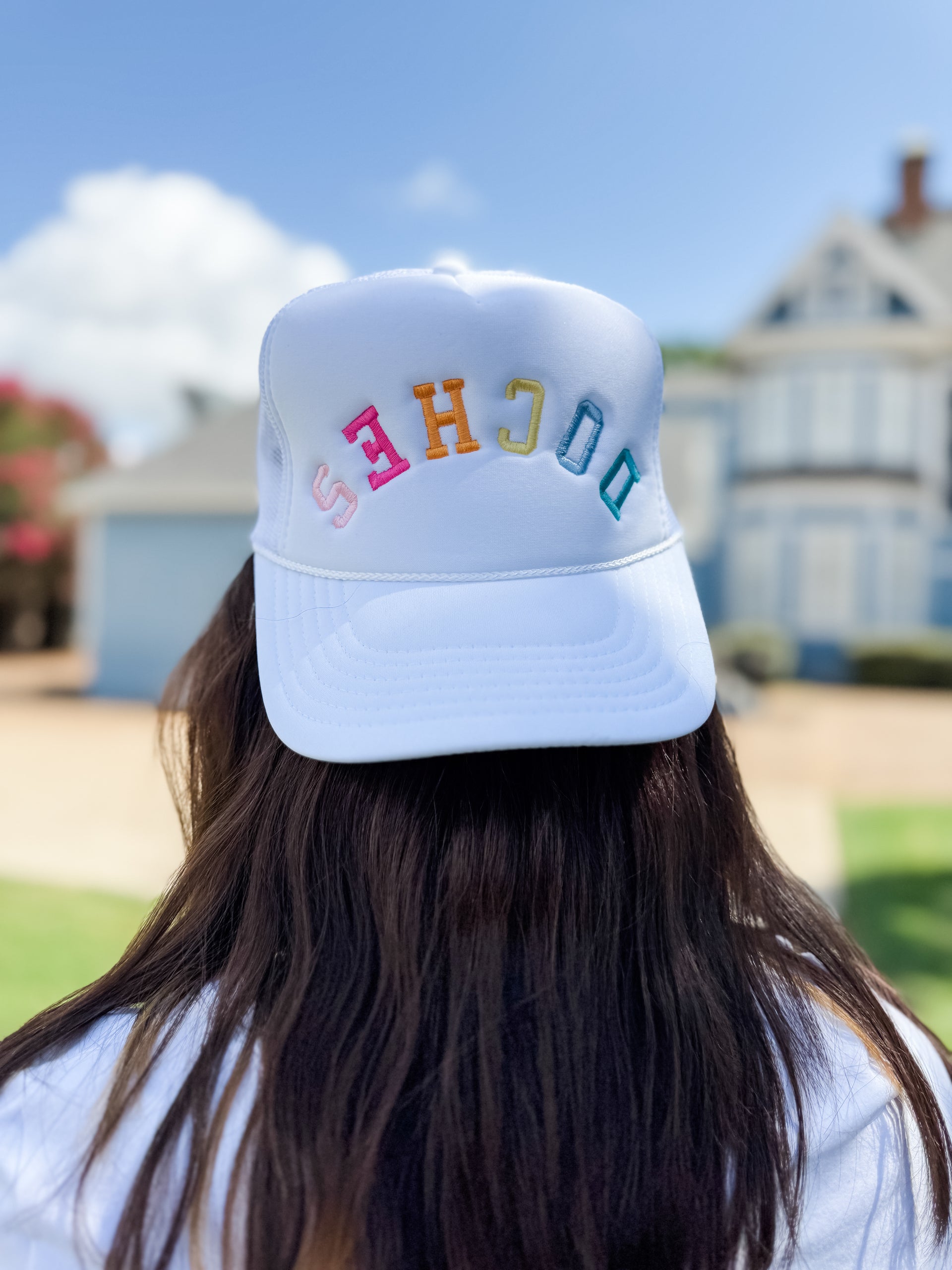 A view from behind of a person with long brown hair wearing a white trucker hat facing backward. The foam front of the hat features the word "DOCHES" embroidered in an arched layout with individual colorful block letters in shades of blue, green, orange, and pink. The background shows a sunny outdoor day with a blurred blue house and green grass.
