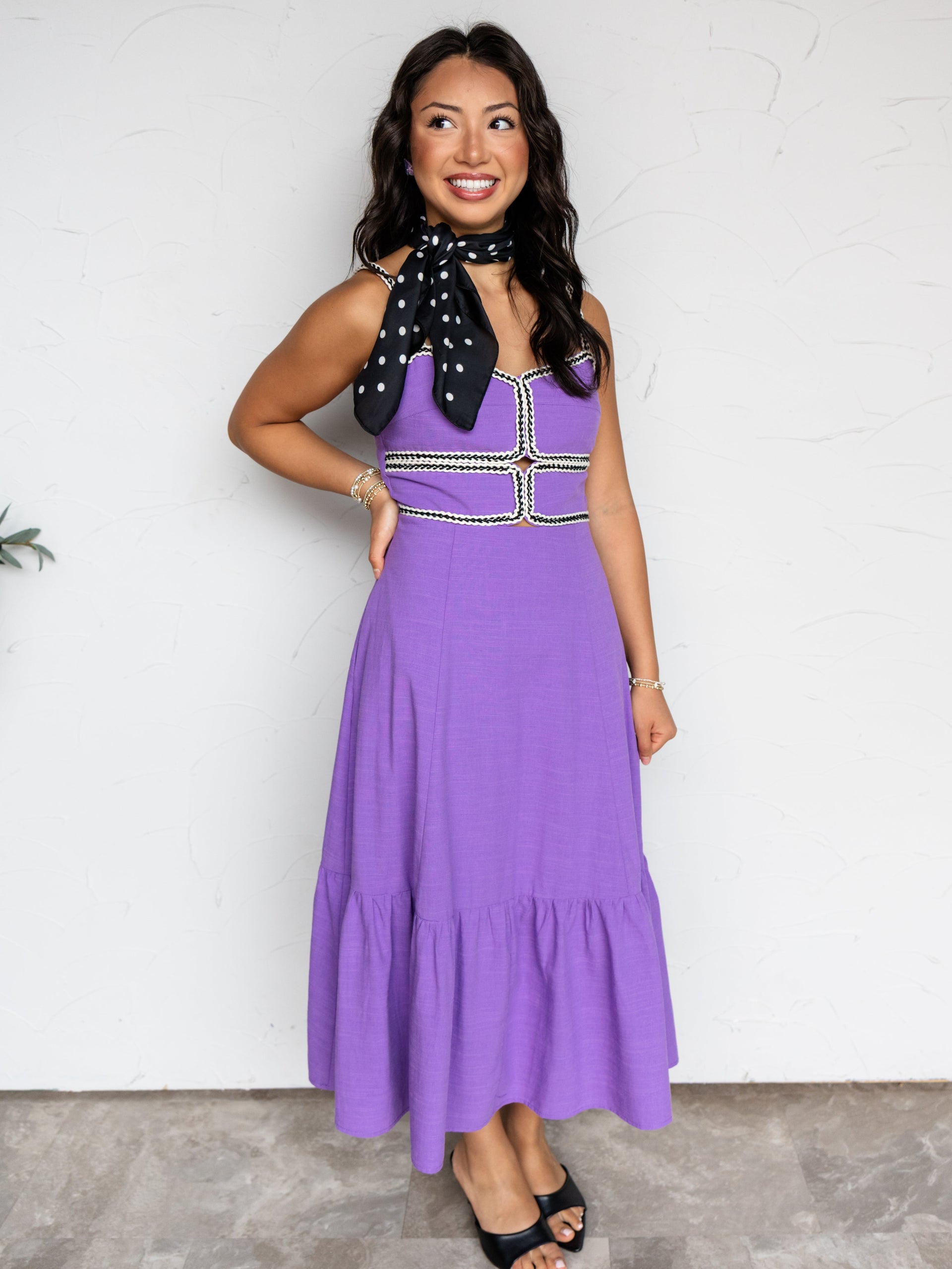 A full-length photograph of a smiling young woman with long dark hair posing against a textured white wall, making a hand gesture. She is wearing a vibrant purple midi dress made from a 65% cotton, 30% viscose, and 5% linen blend fabric, featuring thin cami straps, a bodice accented with black-and-white braided trim and front cutouts, and a tiered ruffle hem, styled with a black-and-white polka dot neck scarf.