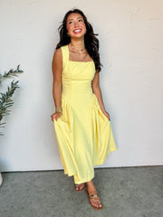 A full-length photograph of a smiling brunette woman looking upward and posing against a textured white wall. She is wearing a pastel yellow midi dress made from a 97% polyester and 3% spandex blend fabric, featuring a square neckline, structured sleeveless bodice, and a gathered drop-waist skirt, styled with flat gold toe-loop sandals.