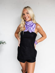 A mid-shot photograph of a smiling blonde woman posing with both hands on her hips against a textured white wall. She is wearing a fitted black strapless romper made from a 70% linen and 30% cotton blend fabric, featuring structural vertical seams along the bodice, styled with a purple paisley patterned neck scarf, purple beaded bracelets, and cowboy boots.
