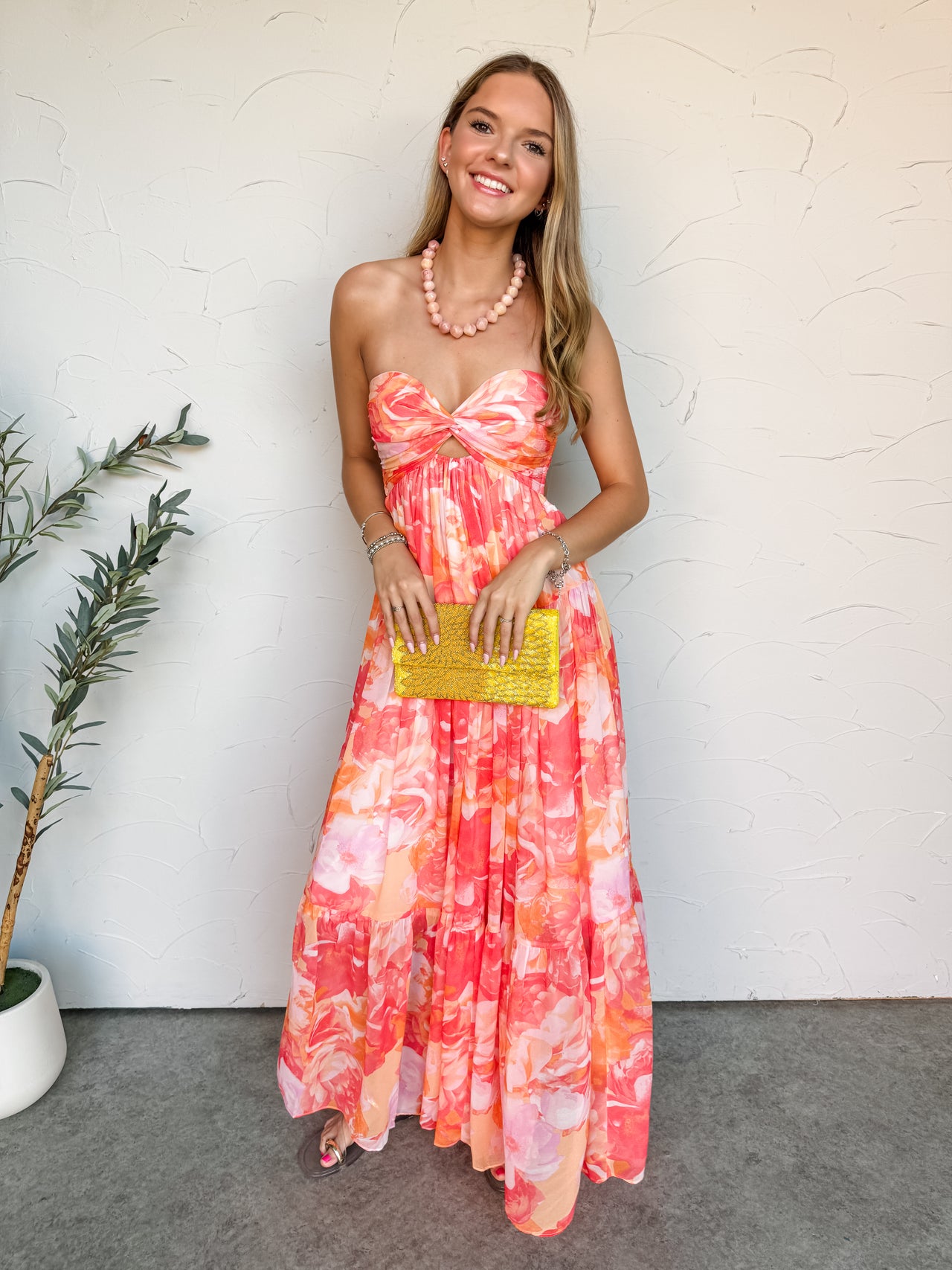 A full-length photograph of a smiling blonde woman posing against a textured white wall. She is wearing a strapless chiffon maxi dress made from 100% polyester fabric in a vibrant coral, pink, and orange watercolor floral print, featuring a twisted sweetheart bust with a small under-bust cutout and a long tiered skirt, accessorized with a chunky pink necklace and a bright yellow beaded clutch bag.
