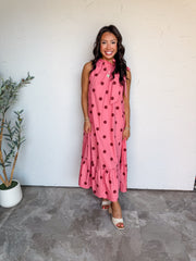 A full-length photograph of a smiling woman with dark wavy hair posing against a textured white wall. She is wearing a sleeveless high-neck maxi dress made from 100% cotton fabric, featuring a bubblegum pink background with large dark reddish-maroon polka dots, a ruffled collar with a side tie bow, and a gathered tiered ruffle hem, paired with off-white open-toe heels and styled with a round gold pendant necklace and gold bracelets.