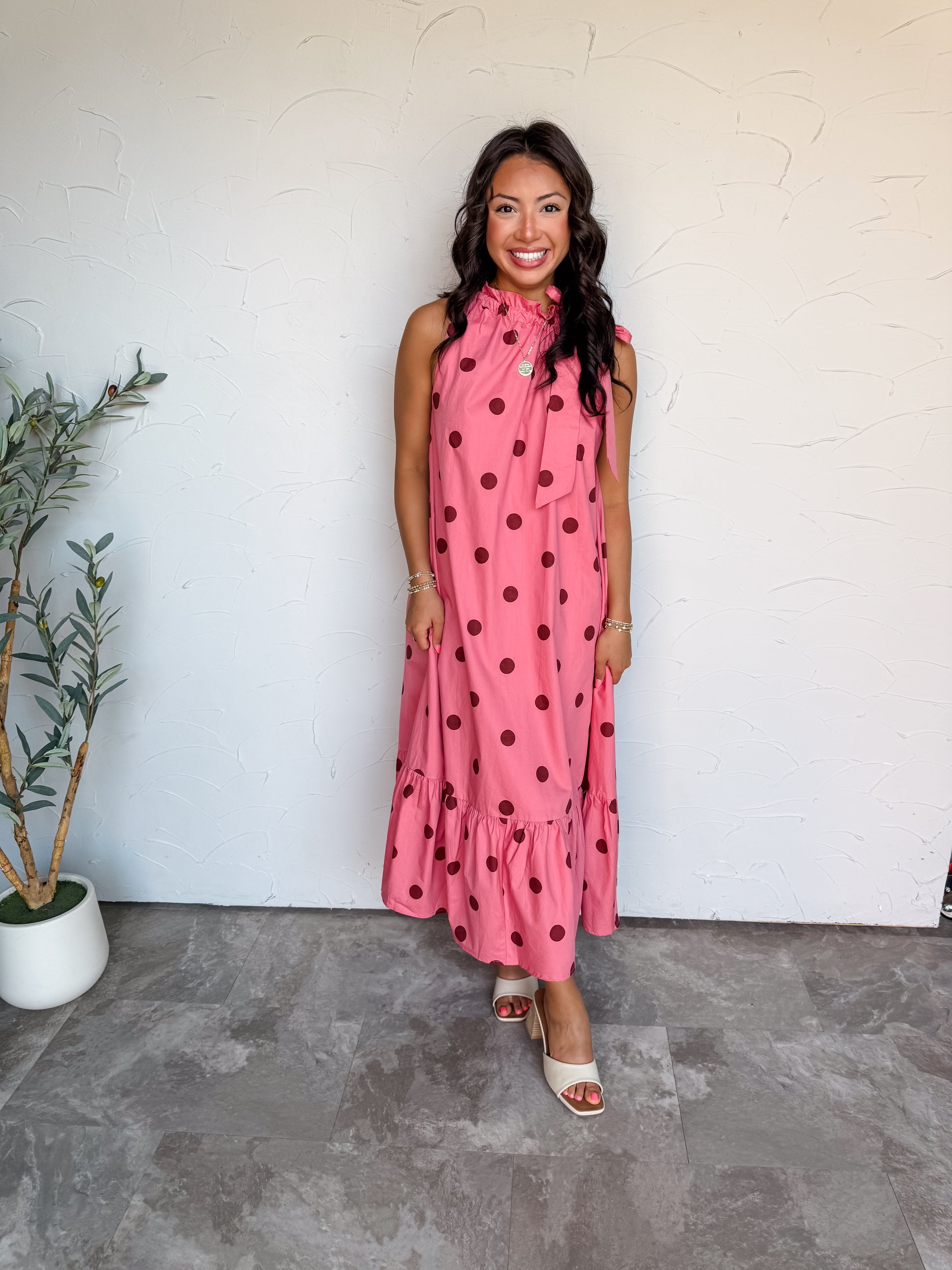 A full-length photograph of a smiling woman with dark wavy hair posing against a textured white wall. She is wearing a sleeveless high-neck maxi dress made from 100% cotton fabric, featuring a bubblegum pink background with large dark reddish-maroon polka dots, a ruffled collar with a side tie bow, and a gathered tiered ruffle hem, paired with off-white open-toe heels and styled with a round gold pendant necklace and gold bracelets.