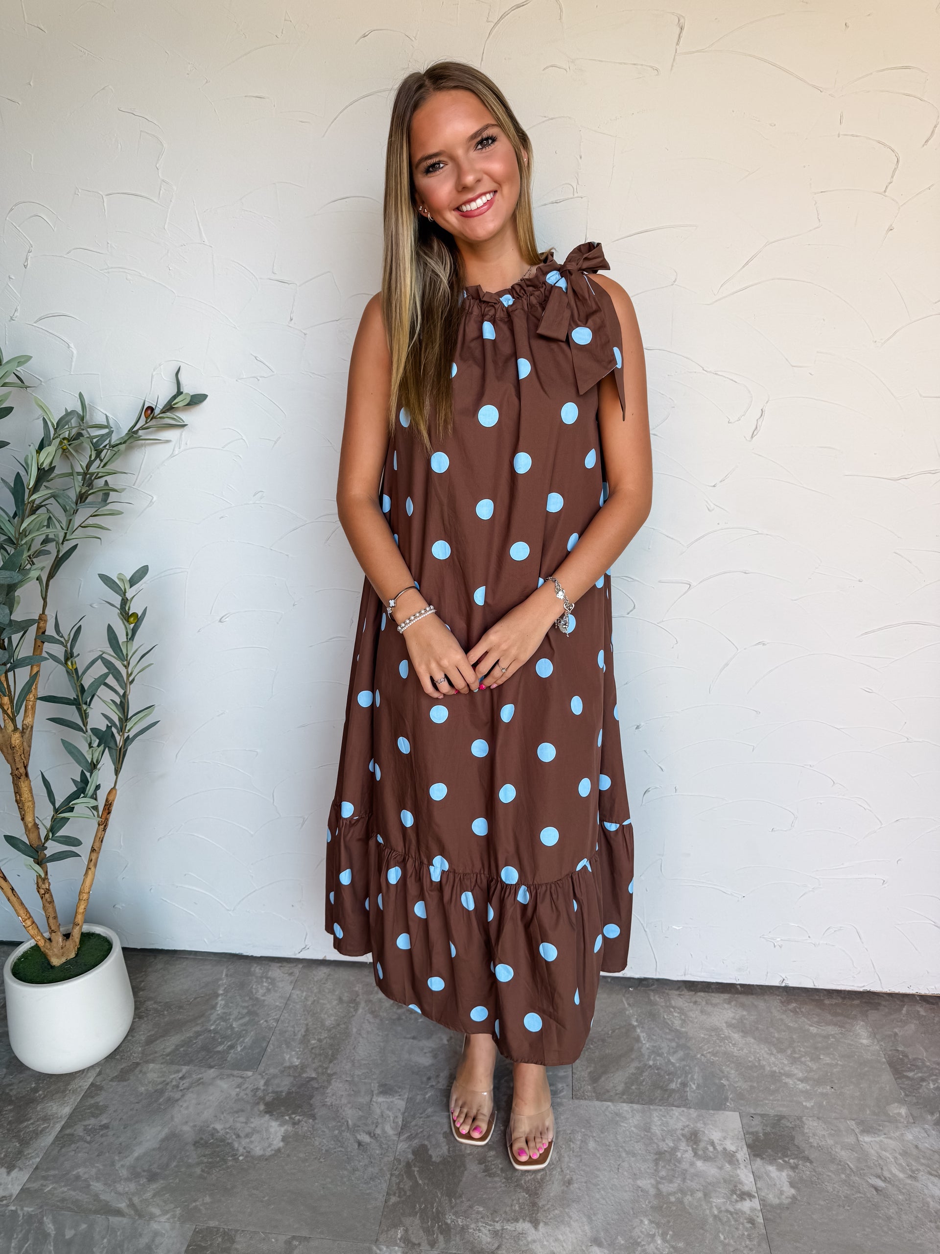 A full-length photograph of a smiling young woman with long blonde-highlighted hair posing against a textured white wall. She is wearing a sleeveless high-neck maxi dress made from 100% cotton fabric, featuring a chocolate brown background with light blue polka dots, a ruffled collar with a bow tie on one shoulder, and a gathered tiered ruffle hem, paired with clear strap sandal heels and delicate silver bracelets.