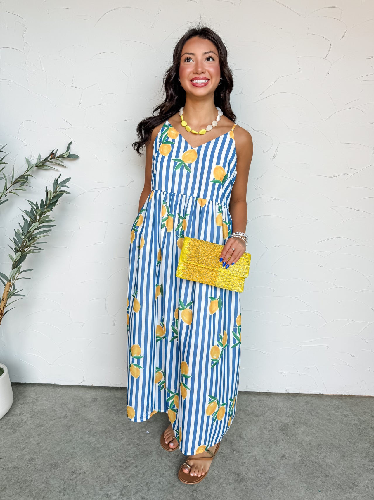 A full-length photograph of a smiling brunette woman posing against a textured white wall. She is wearing a sleeveless maxi dress made from 100% polyester fabric featuring a blue and white vertical stripe pattern covered in bright yellow lemon graphics, designed with thin shoulder straps and a gathered high waist, holding a bright yellow beaded clutch bag.