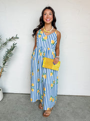 A full-length photograph of a smiling brunette woman posing against a textured white wall. She is wearing a sleeveless maxi dress made from 100% polyester fabric featuring a blue and white vertical stripe pattern covered in bright yellow lemon graphics, designed with thin shoulder straps and a gathered high waist, holding a bright yellow beaded clutch bag.