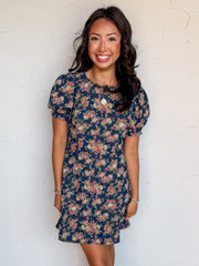 A full-length photograph of a smiling woman with dark wavy hair posing against a textured white wall. She is wearing a short-sleeved mini dress made from an 80% cotton and 20% polyester denim fabric, featuring a dark blue wash covered in a vintage pink and green floral pattern, styled with short puff sleeves, a round neck, and vertical seam details, paired with cream-colored heels and a round gold pendant necklace.