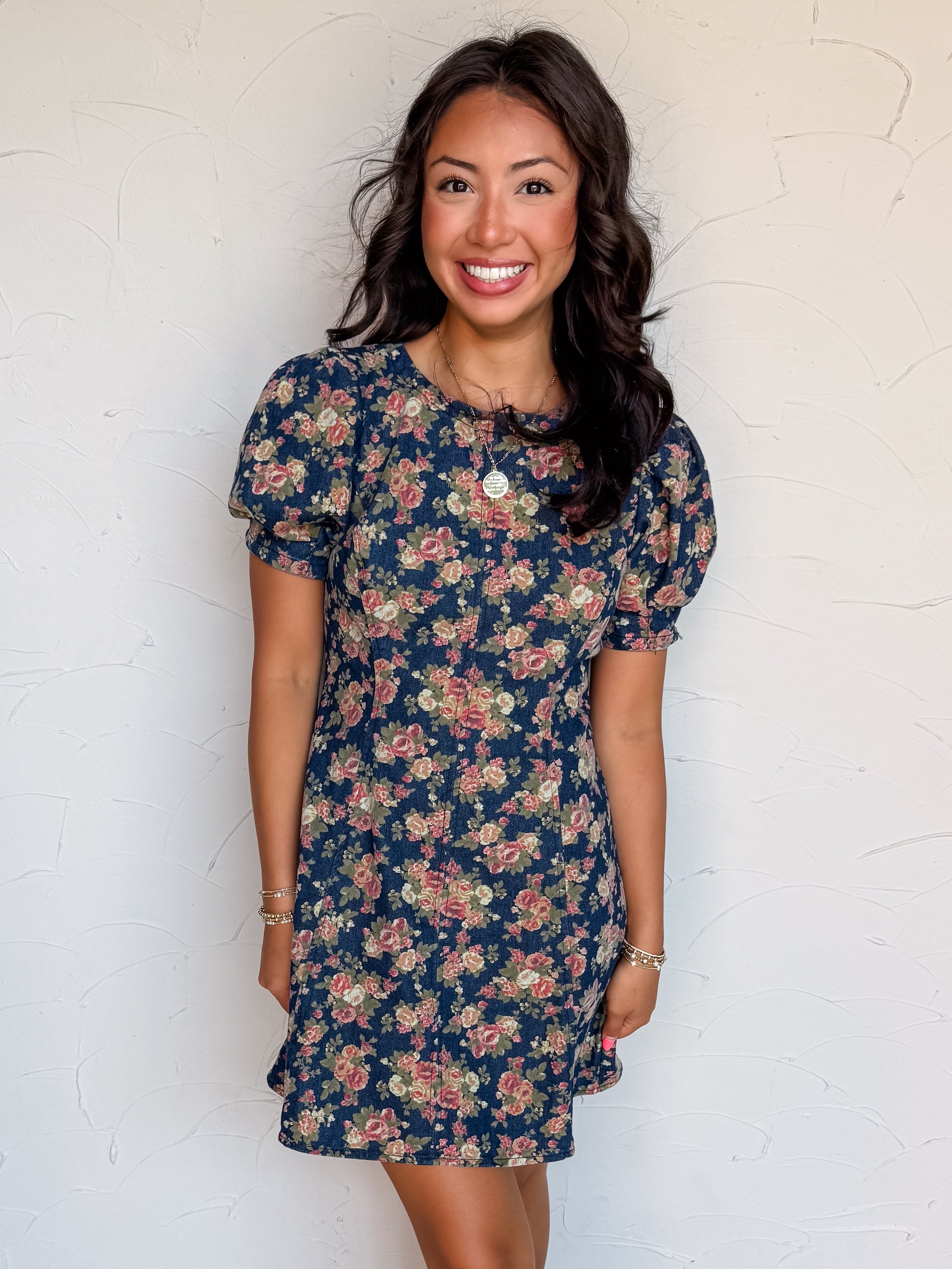 A full-length photograph of a smiling woman with dark wavy hair posing against a textured white wall. She is wearing a short-sleeved mini dress made from an 80% cotton and 20% polyester denim fabric, featuring a dark blue wash covered in a vintage pink and green floral pattern, styled with short puff sleeves, a round neck, and vertical seam details, paired with cream-colored heels and a round gold pendant necklace.