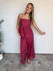 A full-length photograph of a smiling young woman with straight blonde-highlighted hair posing against a textured white wall. She is wearing a two-piece set made from 100% cotton, featuring red and purple stripes; the top is a camisole with thin spaghetti straps, a square neckline, horizontal stripes across the chest, and vertical stripes along the flared peplum waist, paired with matching red and purple vertical striped wide-leg pants, flat gold sandals, and delicate silver and gold necklaces.