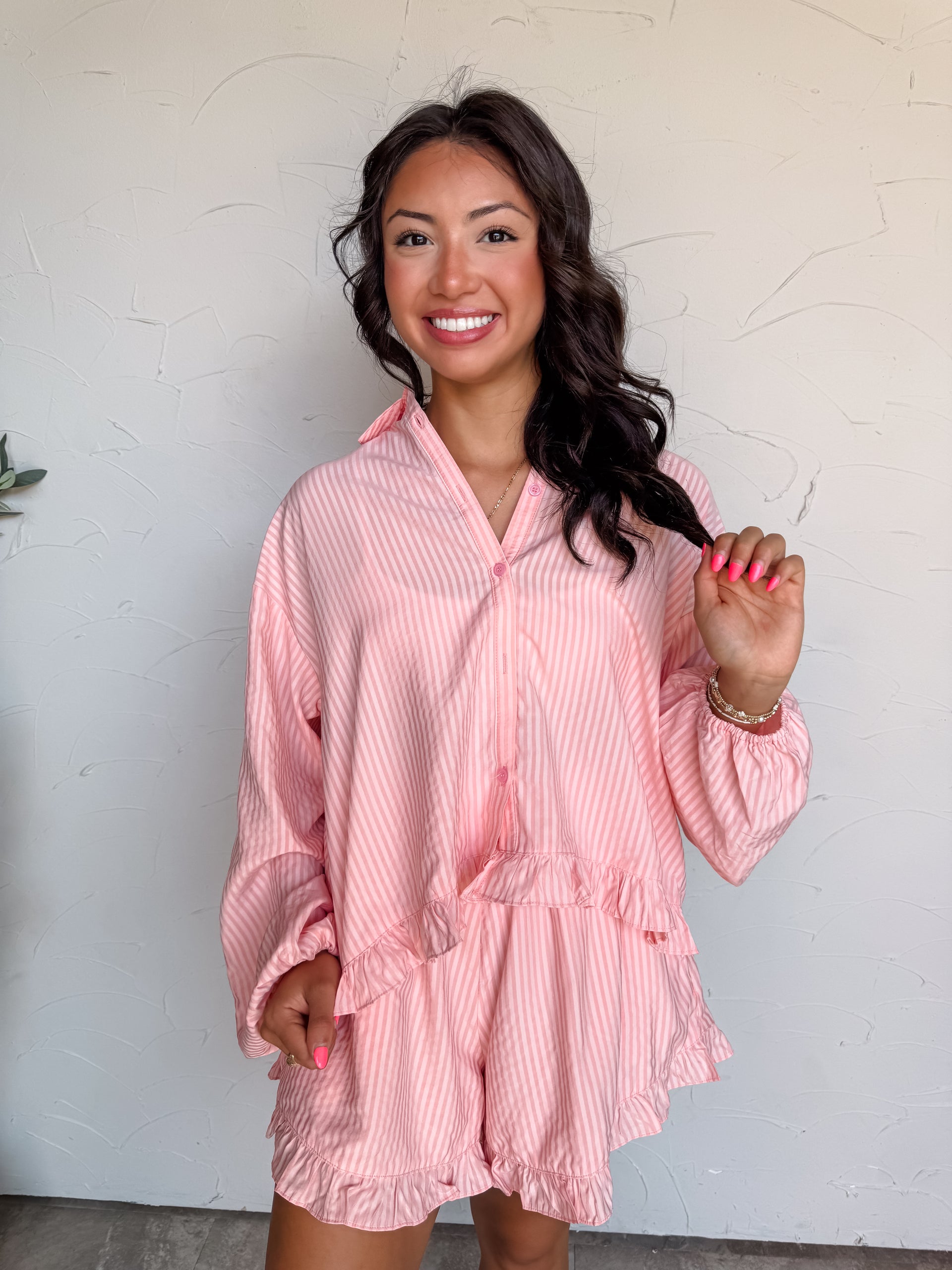 A three-quarter length photograph of an excited woman with dark wavy hair opening her mouth with a playful expression while holding a strand of her hair against a textured white wall. She is wearing a long-sleeved button-down top and matching shorts set made from a 71% rayon, 23% polyester, and 6% nylon blend, featuring fine pink and white vertical stripes, a ruffled hemline, elastic cuffs, paired with matching ruffled striped shorts, and styled with gold chain bracelets and bright pink nail polish.