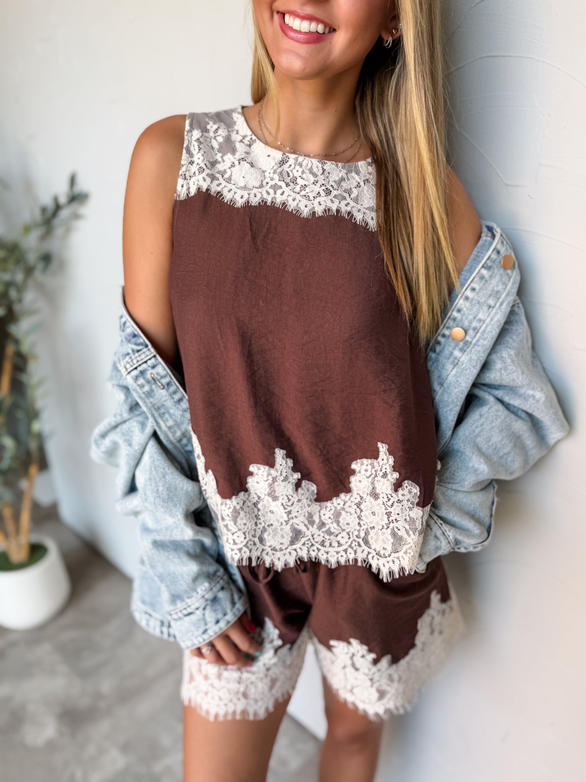 A waist-up photograph of a smiling young woman with straight blonde-highlighted hair posing against a textured white wall. She is wearing a sleeveless chocolate brown top made from an 80% nylon and 20% rayon blend, featuring intricate white eyelash lace trim across the upper neck and bottom hem, paired with matching brown lace-trimmed shorts and an oversized light-wash denim jacket worn casually slouched off her shoulders.