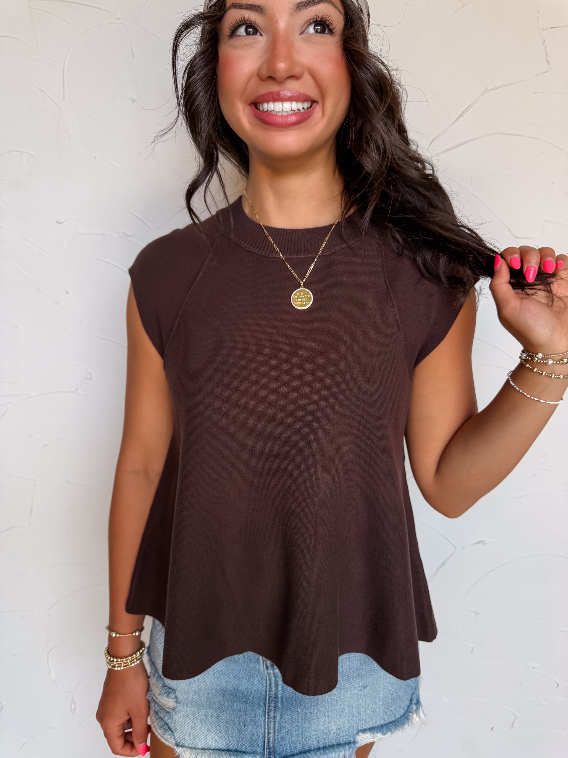 A waist-up photograph of a smiling woman with dark wavy hair looking up towards the side while touching a strand of her hair against a textured white wall. She is wearing a short-sleeved brown knit top made from a 60% viscose, 37% nylon, and 3% spandex blend, featuring cap sleeves, a crew neck, ribbed texture, and a flared peplum hem, styled with a patterned satin headscarf tied around her hair, light-wash frayed denim shorts, a gold pendant necklace, and gold bracelets.
