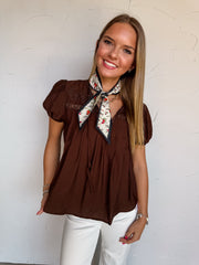 A waist-up photograph of a smiling young woman with straight blonde-highlighted hair posing against a textured white wall. She is wearing a short-sleeved brown blouse made from an 80% rayon and 20% nylon blend, featuring puff sleeves, delicate tonal floral embroidery across the upper chest, a notched split neckline with thin hanging ties, paired with white jeans and styled with a cream floral neck scarf tied at the collar, a gold chain necklace, and silver bracelets.