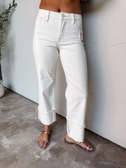 A photograph from the waist down of a model wearing high-waisted white straight-leg jeans against a textured white wall. The jeans are made from a 98% cotton and 2% spandex blend, featuring classic belt loops, five-pocket detail, a front zip fly button closure, built-in tummy control, and wide folded ankle cuffs with raw, frayed hems, paired with flat gold-accented sandals.