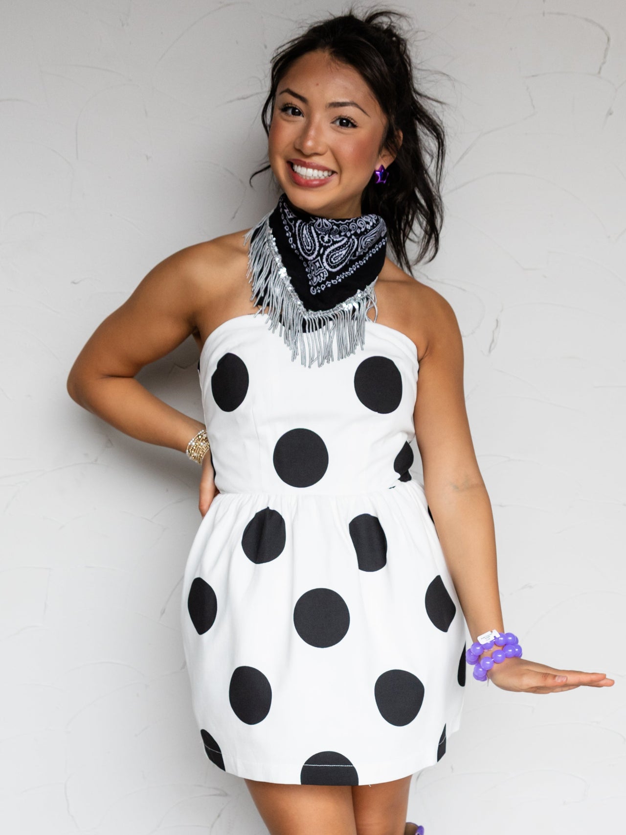 A woman models a white halter mini dress with large black polka dots, styled with a black and white bandana scarf, purple cowboy boots, and a purple wristband against a white wall.