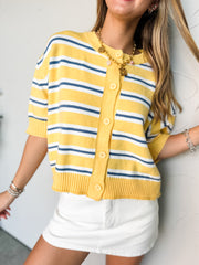 A close-up photograph from the chin down of a blonde woman leaning against a textured white wall. She is wearing a short-sleeve button-front cardigan top made from a 50% viscose, 28% nylon, and 22% polyester blend knit fabric featuring yellow, navy, and white horizontal stripes, styled with matching yellow buttons and high-waisted white denim bottoms.