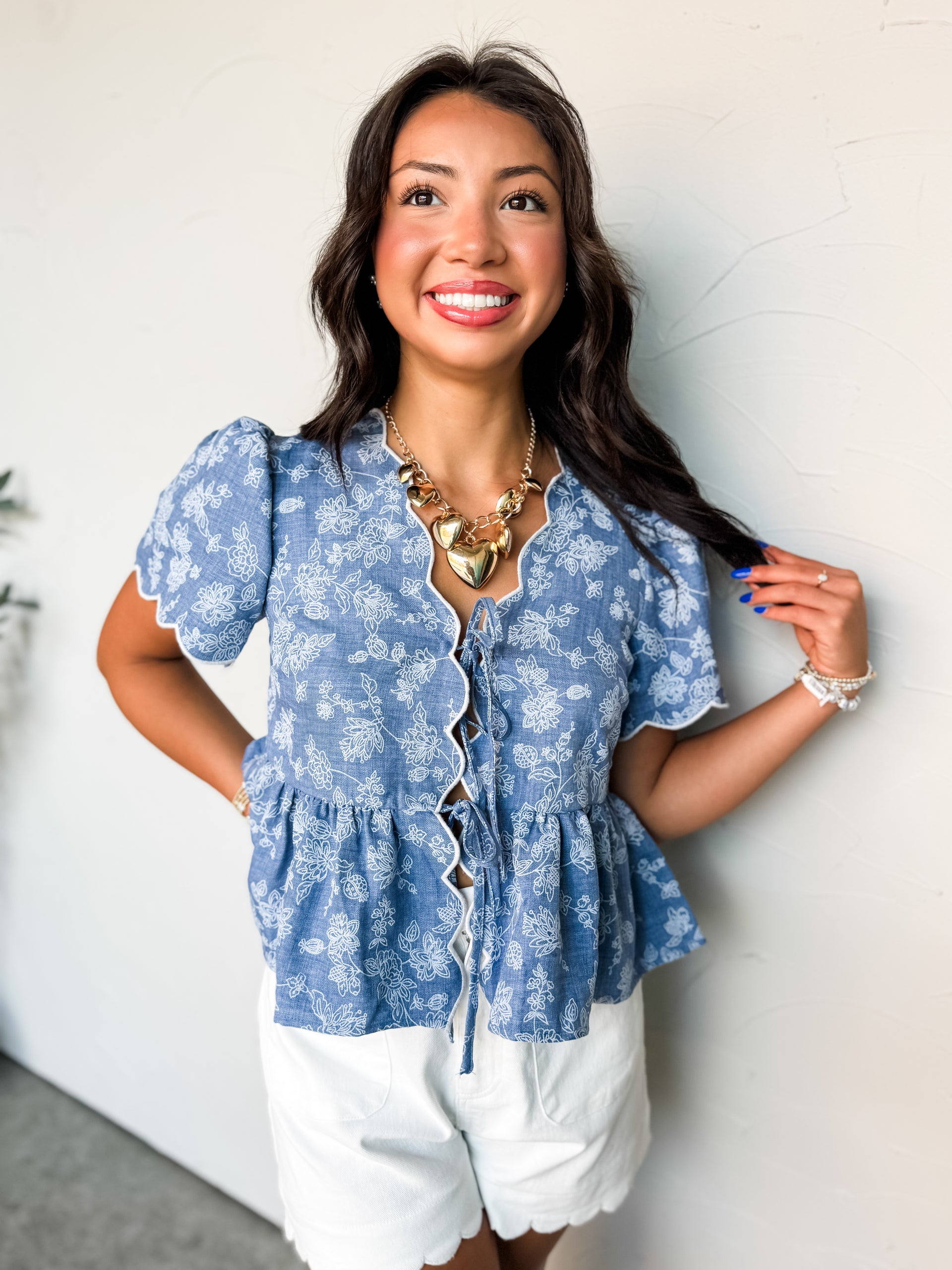 A close-up photograph from the nose down of a brunette woman posing against a textured white wall. She is wearing a short-sleeve chambray-blue top made from 100% polyester fabric featuring a white floral pattern, designed with white scalloped borders along the edges, an open tie-front design, and a peplum hem, styled with white scalloped shorts.