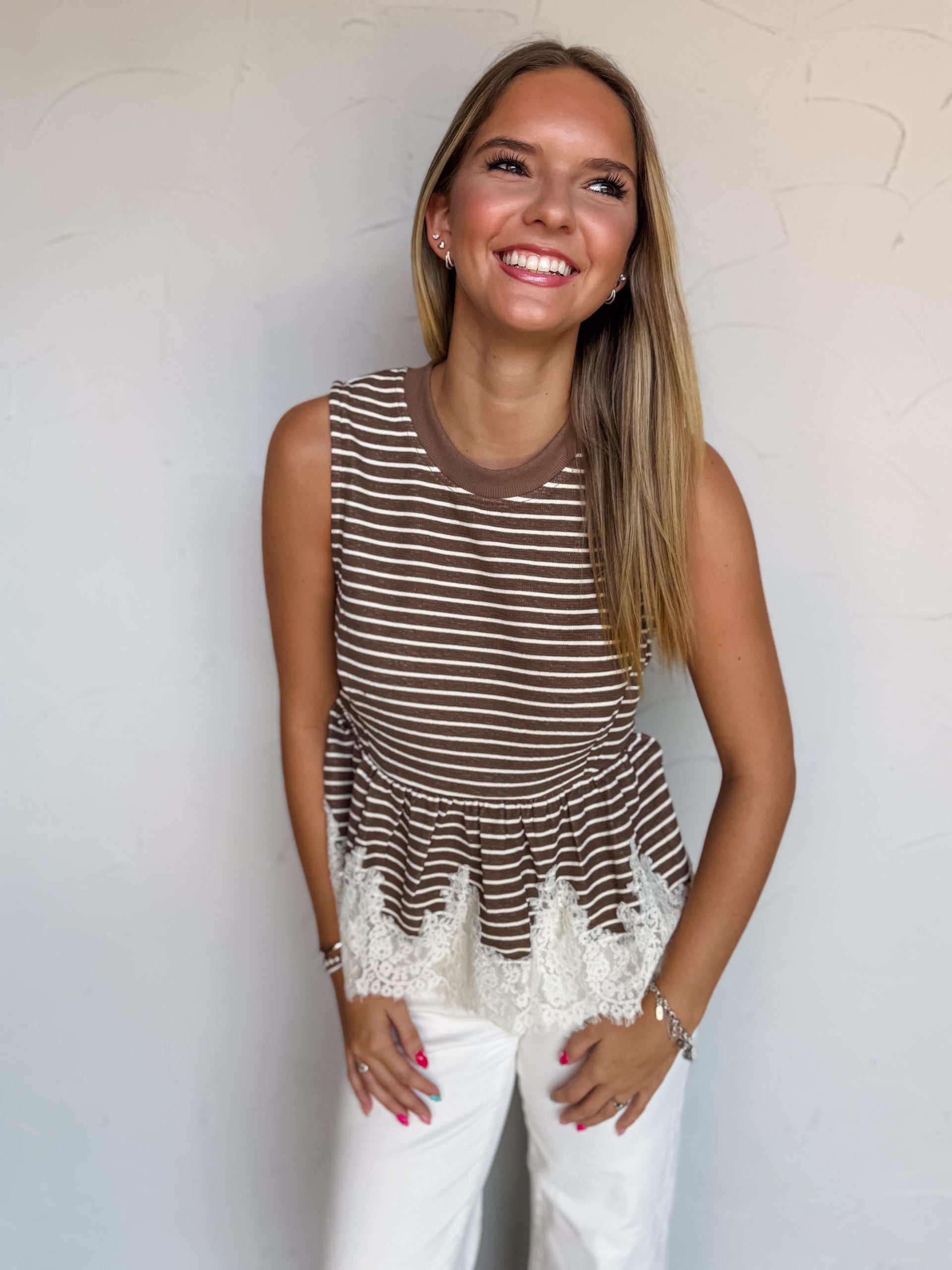 A waist-up photograph of a smiling woman with straight blonde-highlighted hair leaning forward and looking up towards the side against a textured white wall. She is wearing a sleeveless brown and white fine-striped top made from a 97% polyester and 3% spandex blend, featuring a solid brown ribbed crew neckline, a flared peplum waist with delicate white lace trim at the hem, paired with white jeans and styled with silver bracelets.