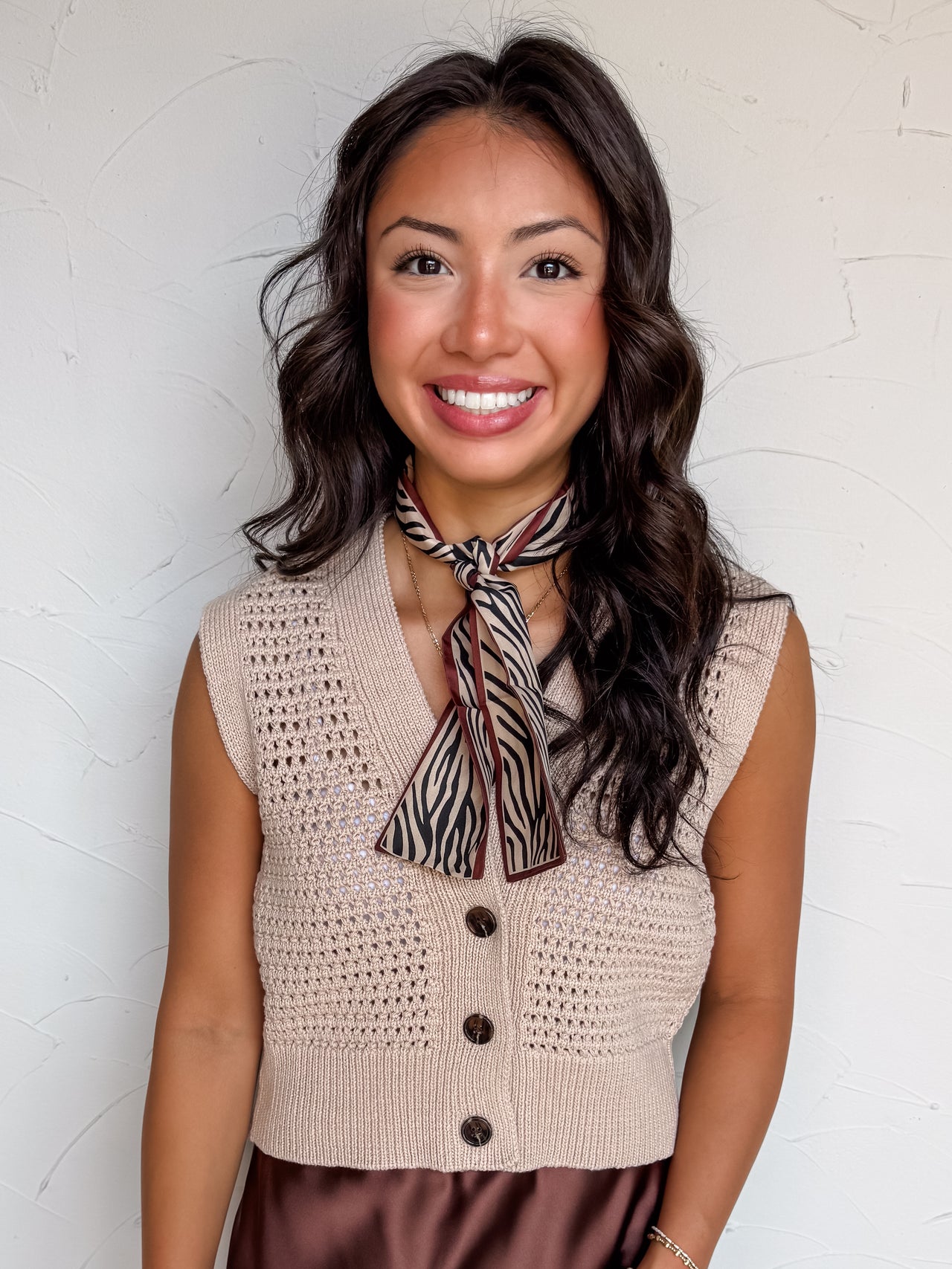 A portrait photograph of a smiling woman with dark wavy hair posing against a textured white wall. She is wearing a light beige sleeveless sweater vest made from 100% cotton, featuring an open pointelle eyelet knit pattern, a button-down front with dark brown tortoiseshell buttons, a V-neckline, and a wide ribbed waistband, styled with a beige and brown zebra-stripe patterned silk neck scarf tied at the collar and a brown satin skirt.