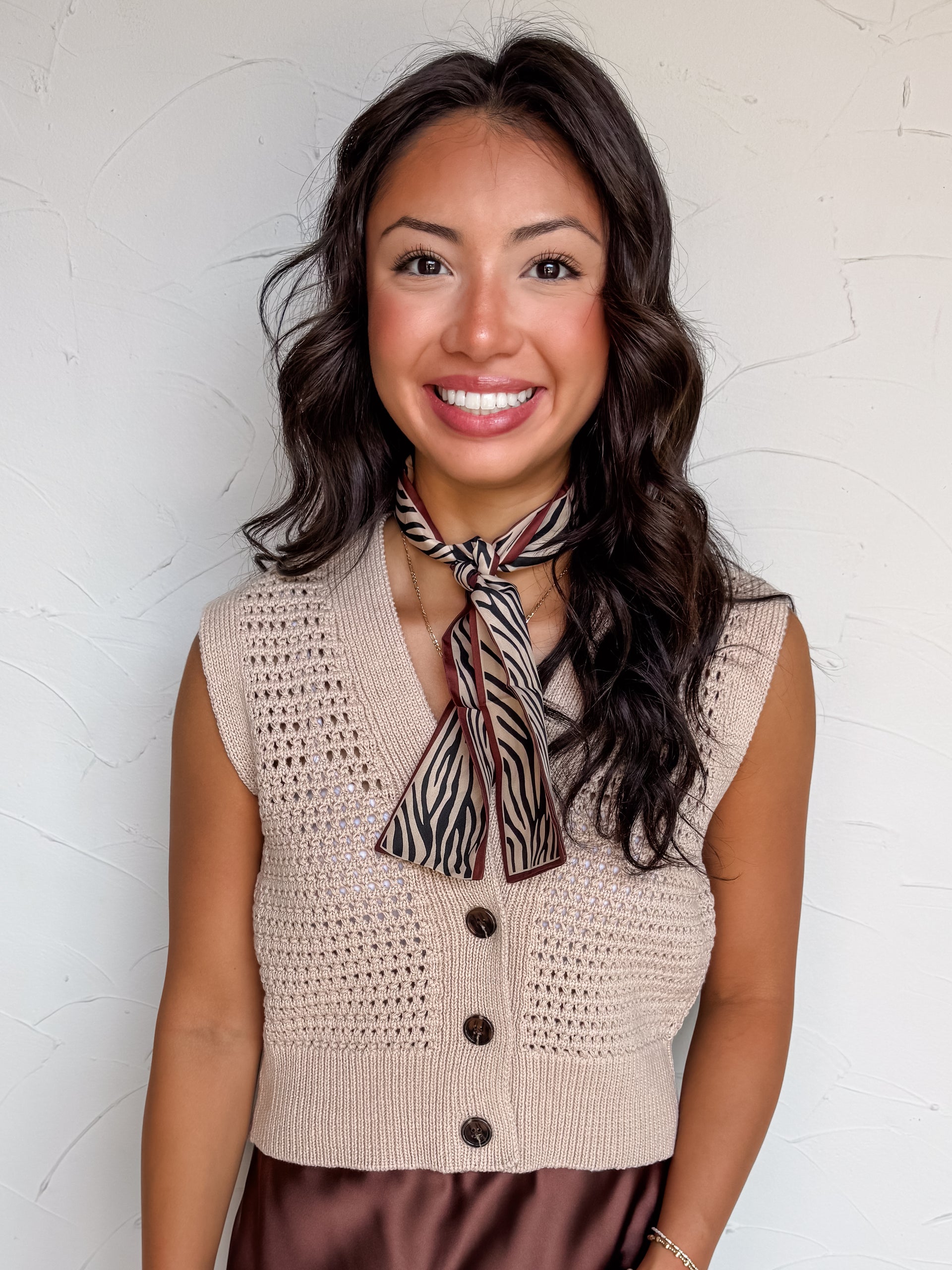 A portrait photograph of a smiling woman with dark wavy hair posing against a textured white wall. She is wearing a light beige sleeveless sweater vest made from 100% cotton, featuring an open pointelle eyelet knit pattern, a button-down front with dark brown tortoiseshell buttons, a V-neckline, and a wide ribbed waistband, styled with a beige and brown zebra-stripe patterned silk neck scarf tied at the collar and a brown satin skirt.