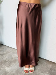 A photograph from the waist down of a model wearing a high-waisted brown satin midi skirt against a textured white wall. The skirt is made from 100% polyester fabric, featuring a smooth chocolate brown finish with a subtle sheen, a comfortable elastic waist, and a fluid bias-cut hem reaching the lower calves, paired with cream open-toe slide heels.
