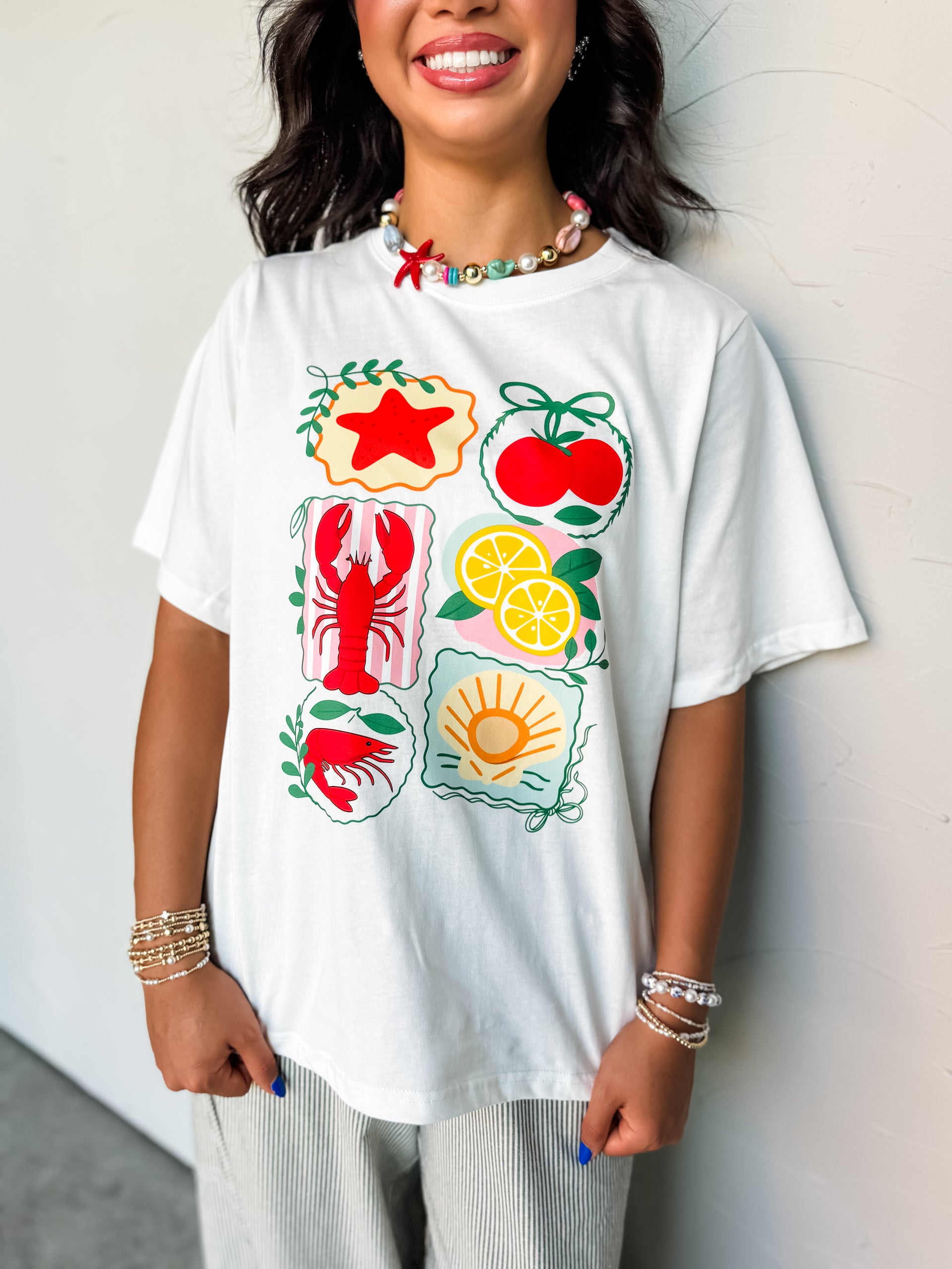 A close-up photograph from the nose down of a smiling brunette woman posing against a textured white wall. She is wearing a relaxed white short-sleeve t-shirt made from 100% cotton fabric featuring a colorful graphic print with individual illustrated panels of seafood and fruits, styled over green and white vertically striped pants.