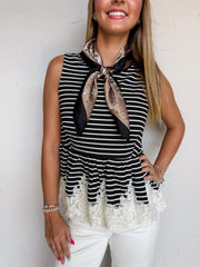 A waist-up photograph of a smiling woman with straight blonde-highlighted hair posing with one hand on her hip against a textured white wall. She is wearing a sleeveless black and white fine-striped top made from a 97% polyester and 3% spandex blend, featuring a flared peplum waist with delicate white lace trim at the hem, styled with a bronze and black silk paisley neck scarf tied at the collar, white jeans, and silver bracelets.