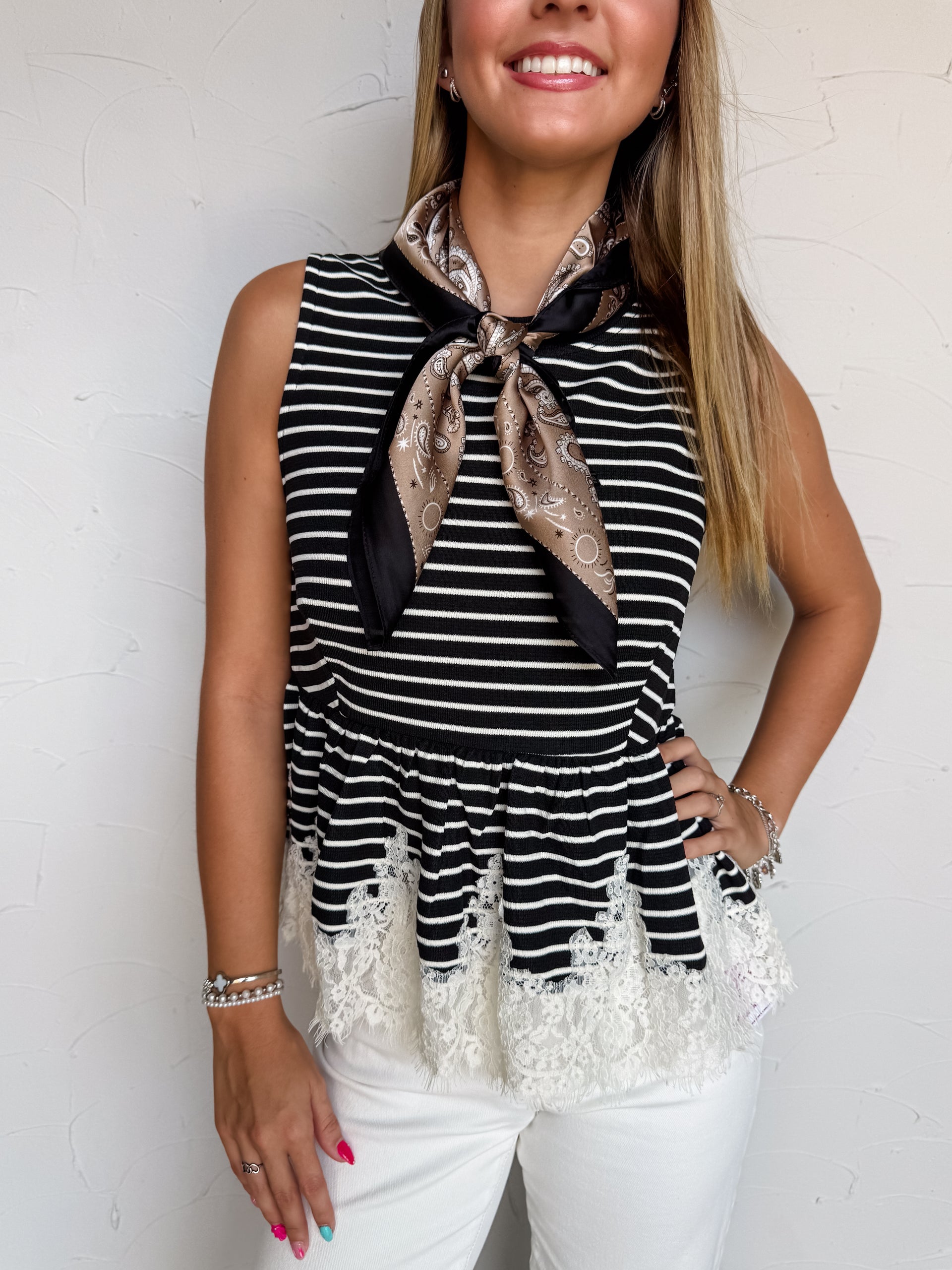 A waist-up photograph of a smiling woman with straight blonde-highlighted hair posing with one hand on her hip against a textured white wall. She is wearing a sleeveless black and white fine-striped top made from a 97% polyester and 3% spandex blend, featuring a flared peplum waist with delicate white lace trim at the hem, styled with a bronze and black silk paisley neck scarf tied at the collar, white jeans, and silver bracelets.