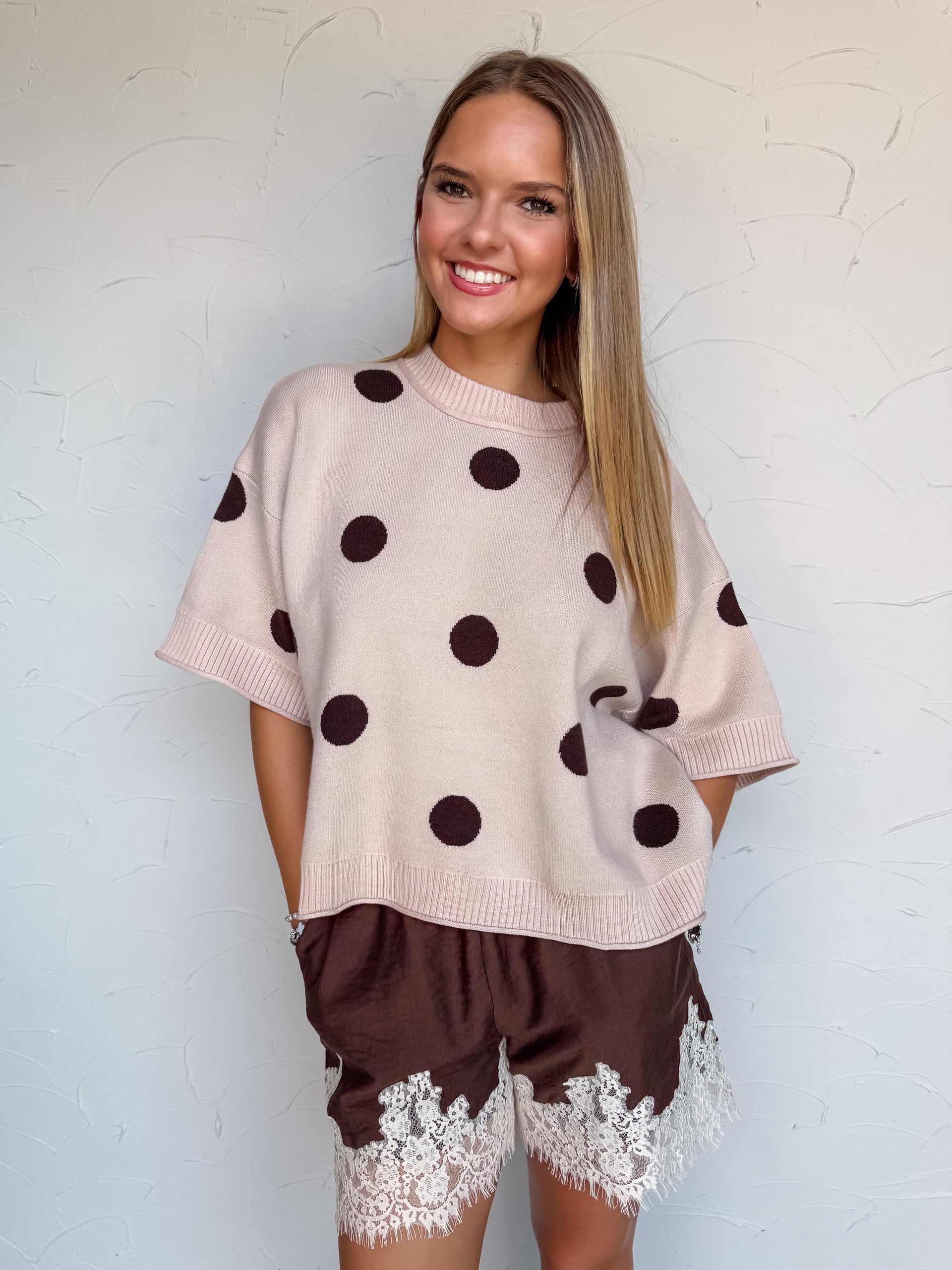 A full-length photograph of a smiling young woman with straight blonde-highlighted hair posing dynamically on one leg with arms outstretched against a textured white wall. She is wearing a soft beige short-sleeved knit sweater made from a 48% viscose, 28% polyester, and 24% nylon blend, featuring large dark brown polka dots, ribbed trim, and wide sleeves, front-tucked into high-waisted dark brown shorts with white lace trim, paired with brown flats and styled with silver chain bracelets.