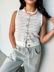 A close-up photograph from the nose down of a brunette woman leaning against a textured white wall. She is wearing a sleeveless knit vest made from a 65% acrylic and 35% nylon blend fabric featuring colorful horizontal stripes, styled with a crew neckline and a button-down front with round wooden buttons, tucked slightly into grey and white striped drawstring pants.