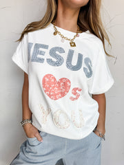 A close-up photograph from the chin down of a blonde woman posing with her hands in her pockets against a textured white wall. She is wearing a relaxed white short-sleeve sweatshirt made from an 85% cotton and 15% polyester blend fabric, featuring printed patchwork lettering and a floral heart pattern that spells out "Jesus Loves You," styled over blue and white vertically striped drawstring pants.