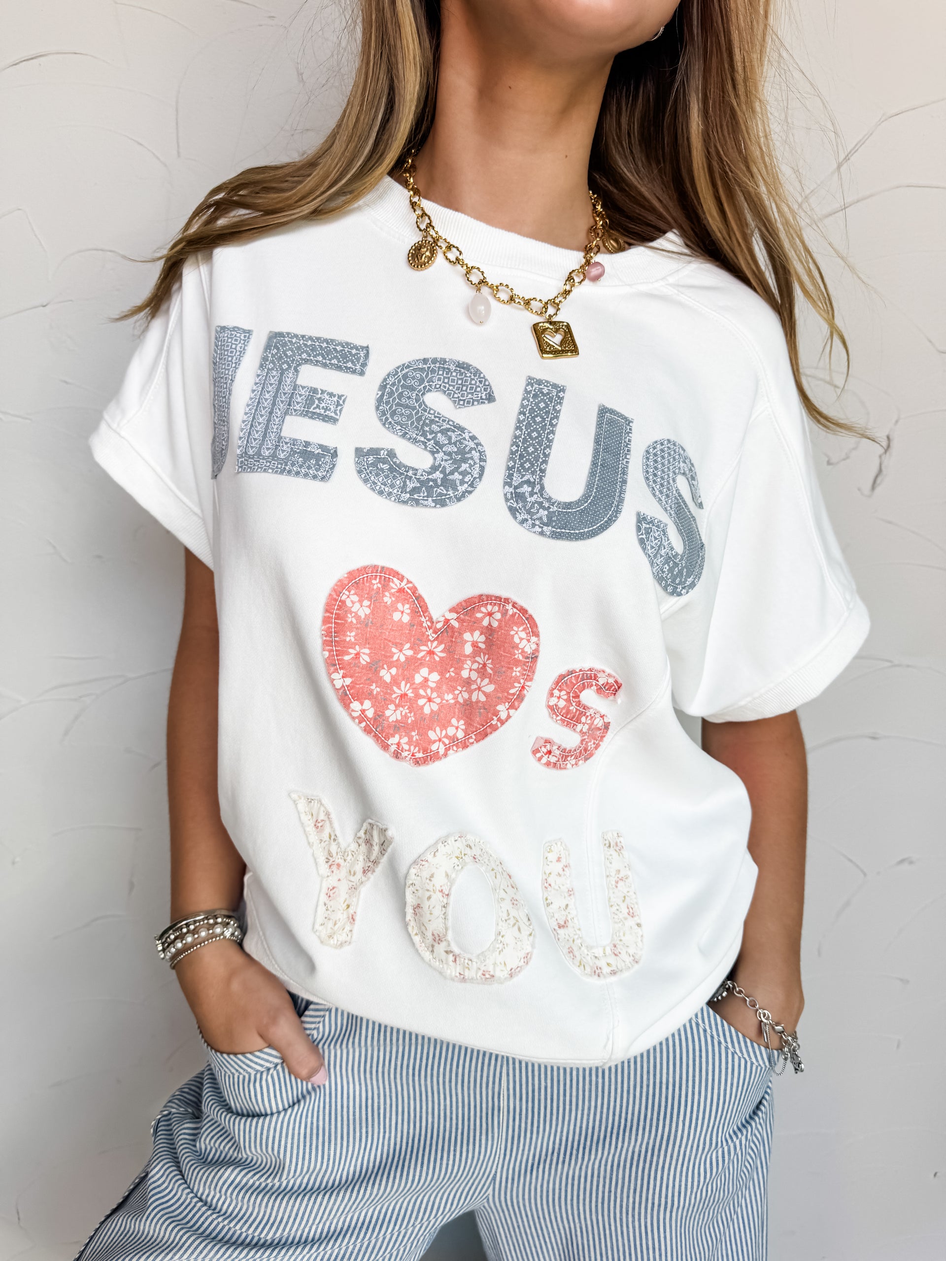 A close-up photograph from the chin down of a blonde woman posing with her hands in her pockets against a textured white wall. She is wearing a relaxed white short-sleeve sweatshirt made from an 85% cotton and 15% polyester blend fabric, featuring printed patchwork lettering and a floral heart pattern that spells out "Jesus Loves You," styled over blue and white vertically striped drawstring pants.