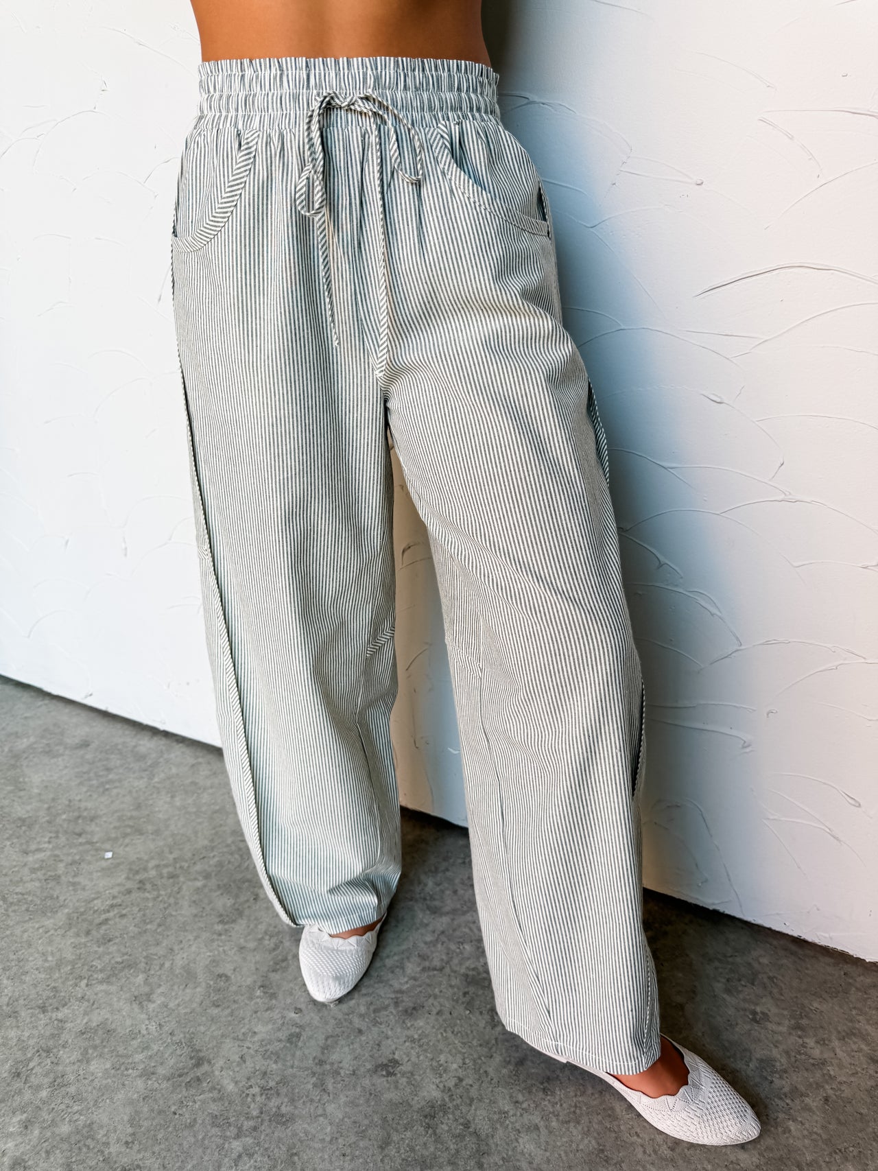 A close-up photograph focusing from the waist down of a woman leaning against a textured white wall. She is wearing high-waisted, wide-leg pants made from 100% cotton fabric featuring a sage green and white vertical stripe pattern, styled with an elastic drawstring waistband and deep front scoop pockets, showing her white woven flat shoes at the bottom.