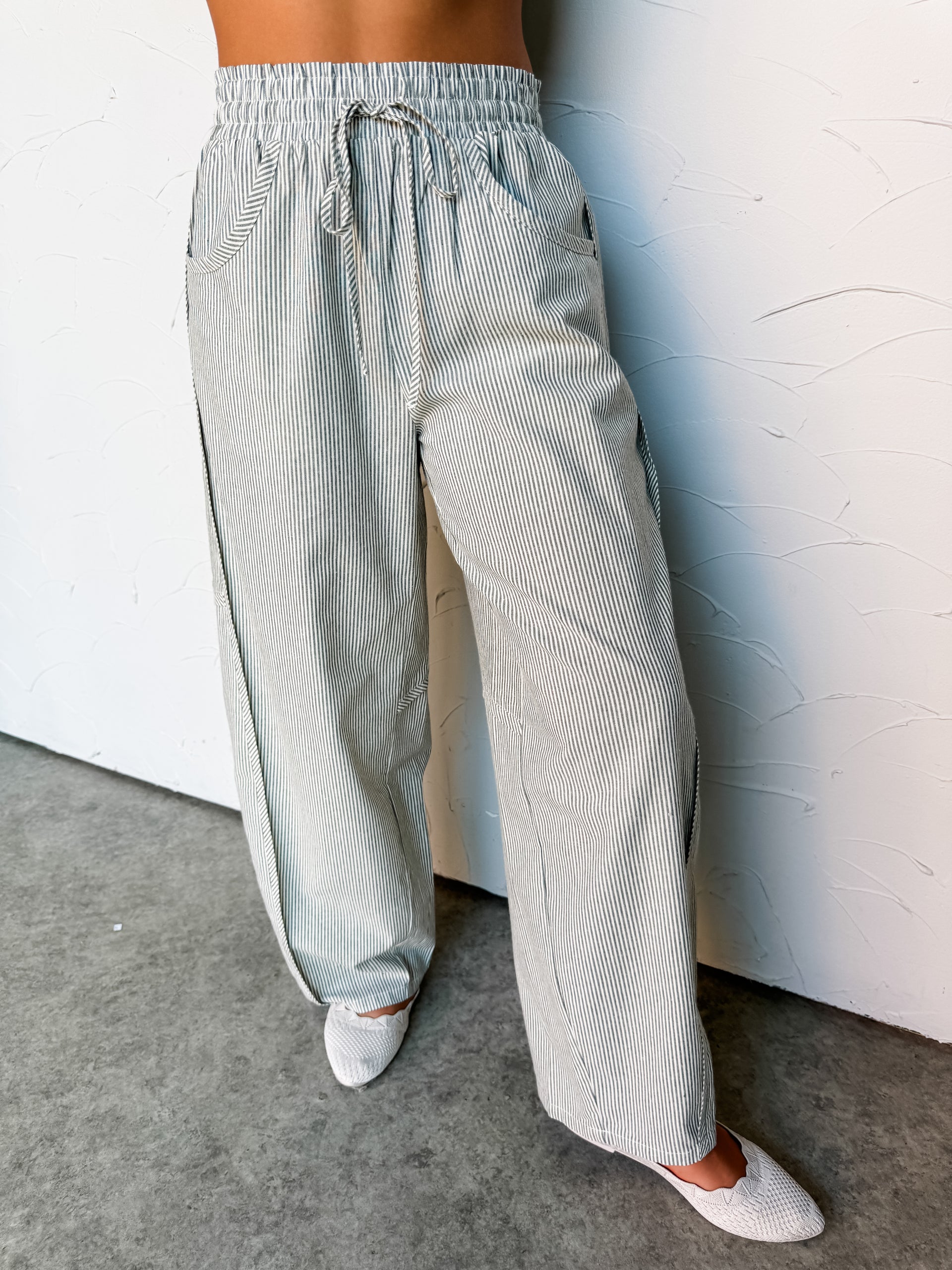A close-up photograph focusing from the waist down of a woman leaning against a textured white wall. She is wearing high-waisted, wide-leg pants made from 100% cotton fabric featuring a sage green and white vertical stripe pattern, styled with an elastic drawstring waistband and deep front scoop pockets, showing her white woven flat shoes at the bottom.