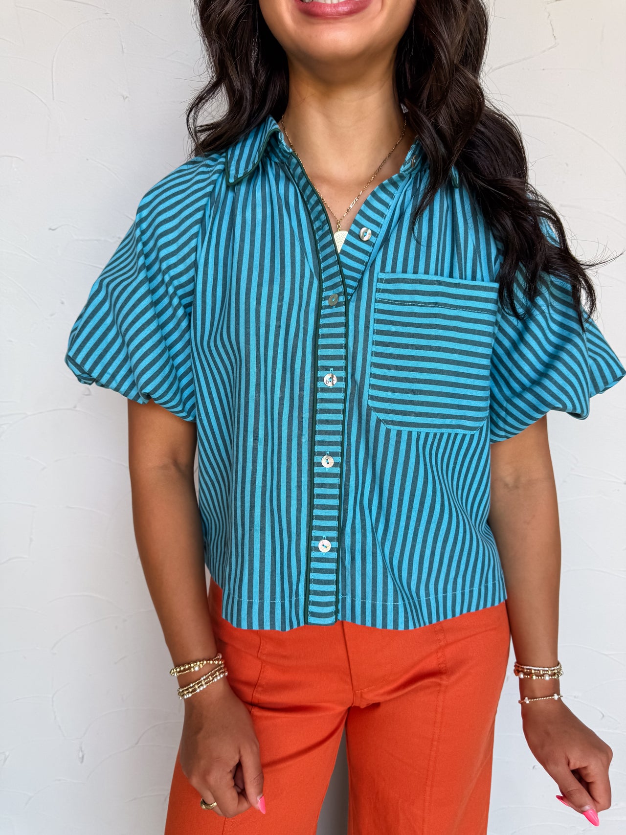 A waist-up photograph of a smiling woman with dark wavy hair posing against a textured white wall. She is wearing a short-sleeved button-down top made from a 50% cotton and 50% polyester blend, featuring teal blue and dark green vertical stripes, light-colored front buttons, wide loose sleeves, and a patch chest pocket with horizontal stripes, tucked into vibrant orange wide-leg pants and styled with a gold pendant necklace and gold beaded bracelets.