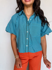 A waist-up photograph of a smiling woman with dark wavy hair posing against a textured white wall. She is wearing a short-sleeved button-down top made from a 50% cotton and 50% polyester blend, featuring teal blue and dark green vertical stripes, light-colored front buttons, wide loose sleeves, and a patch chest pocket with horizontal stripes, tucked into vibrant orange wide-leg pants and styled with a gold pendant necklace and gold beaded bracelets.
