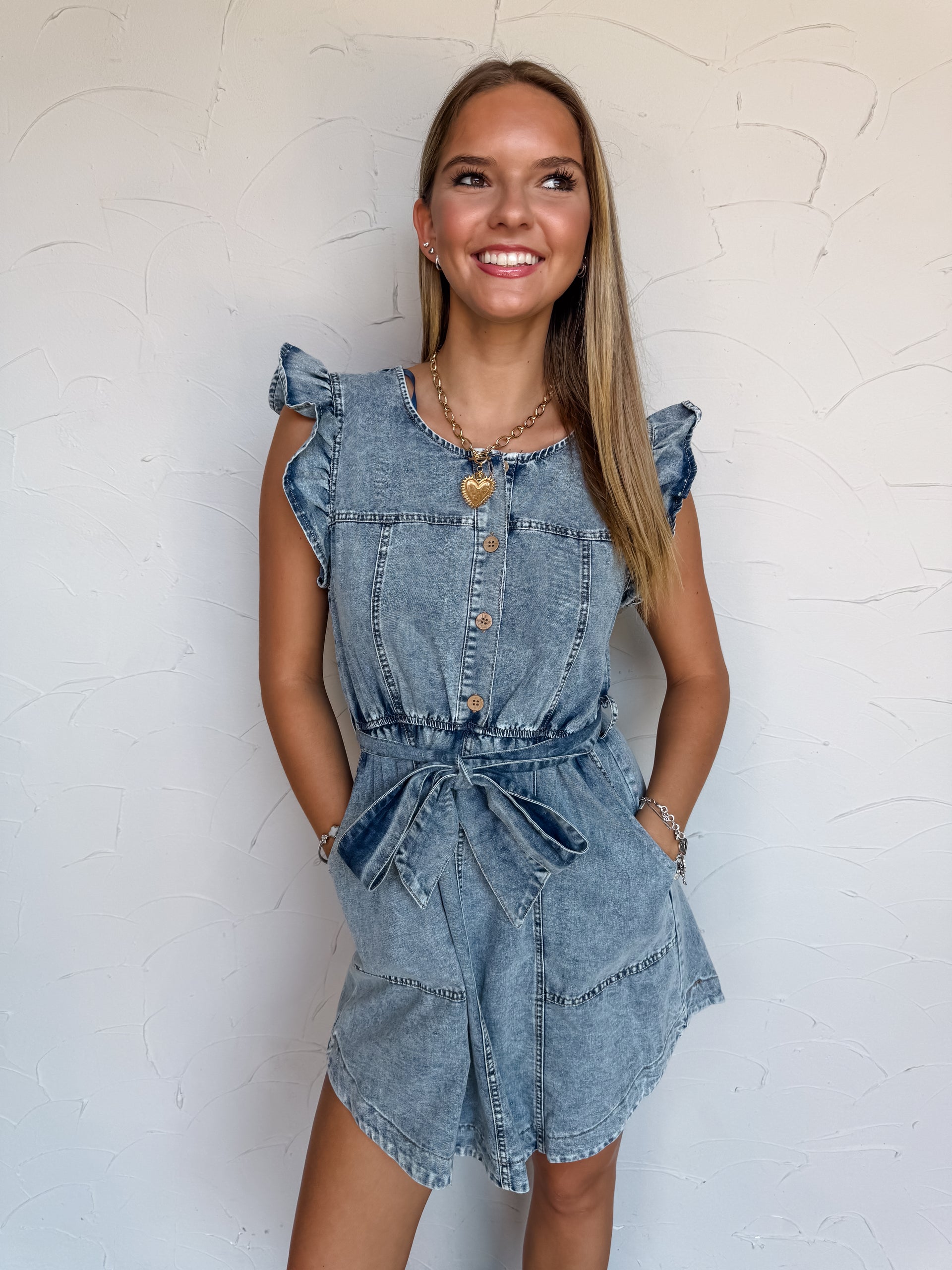 A waist-up photograph of a smiling young woman with long blonde-highlighted hair looking off-camera against a textured white wall with hands in her pockets. She is wearing a medium-wash blue denim romper made from 100% cotton, featuring flutter ruffle trim at the shoulders, a round neckline, three front buttons, a belted waist tied in a bow, and front pockets, accessorized with a thick gold chain necklace with a heart pendant.