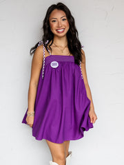 A full-length photograph of a smiling young woman with long dark wavy hair posing against a textured white wall. She is wearing a relaxed purple mini dress made from 100% cotton fabric, featuring a smocked bust line and braided purple-and-white shoulder straps that tie at the shoulders, styled with a round purple pin on the chest, tall white western cowboy boots, a gold chain necklace, and stacked gold bracelets.