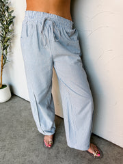 A close-up photograph focusing from the waist down of a woman leaning against a textured white wall. She is wearing high-waisted, wide-leg pants made from 100% cotton fabric featuring a blue and white vertical stripe pattern, styled with an elastic drawstring waistband and deep front scoop pockets, showing her sandaled feet at the bottom.

