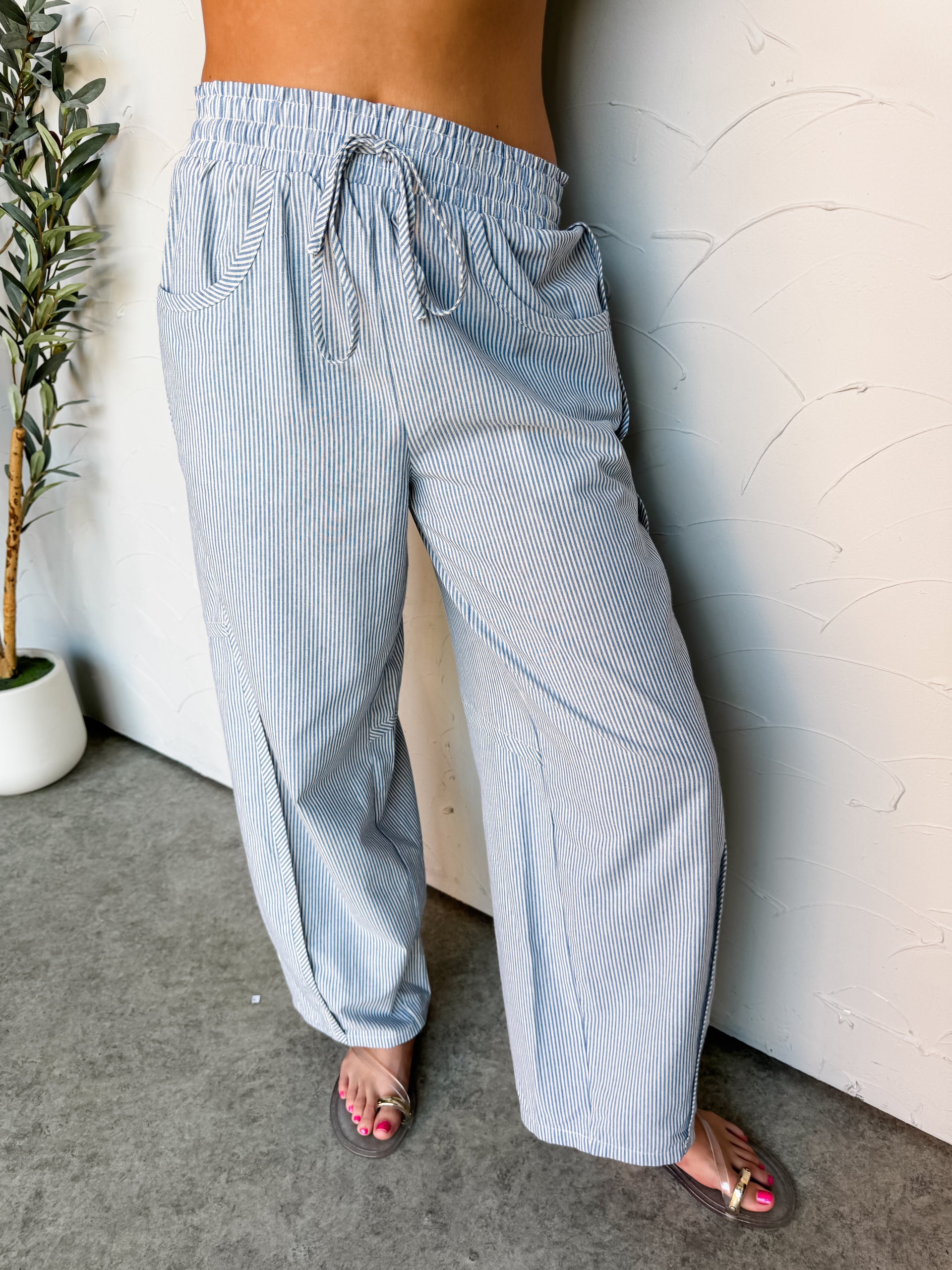 A close-up photograph focusing from the waist down of a woman leaning against a textured white wall. She is wearing high-waisted, wide-leg pants made from 100% cotton fabric featuring a blue and white vertical stripe pattern, styled with an elastic drawstring waistband and deep front scoop pockets, showing her sandaled feet at the bottom.

