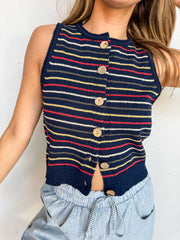 A close-up photograph from the nose down of a blonde woman leaning against a textured white wall. She is wearing a sleeveless knit vest made from a 65% acrylic and 35% nylon blend fabric featuring a navy base with red, yellow, and white horizontal stripes, styled with a crew neckline and a button-down front with round wooden buttons, over blue and white striped drawstring pants.