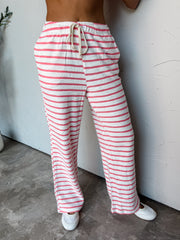 A photograph from the waist down of a model wearing high-waisted striped lounge pants against a textured white wall. The pants are made from an 87% cotton and 13% polyester blend, featuring a white textured knit base with vibrant pink horizontal stripes, an elastic waistband with a thick cream braided rope drawstring tie, and a full-length wide-leg cut, paired with white sneakers.
