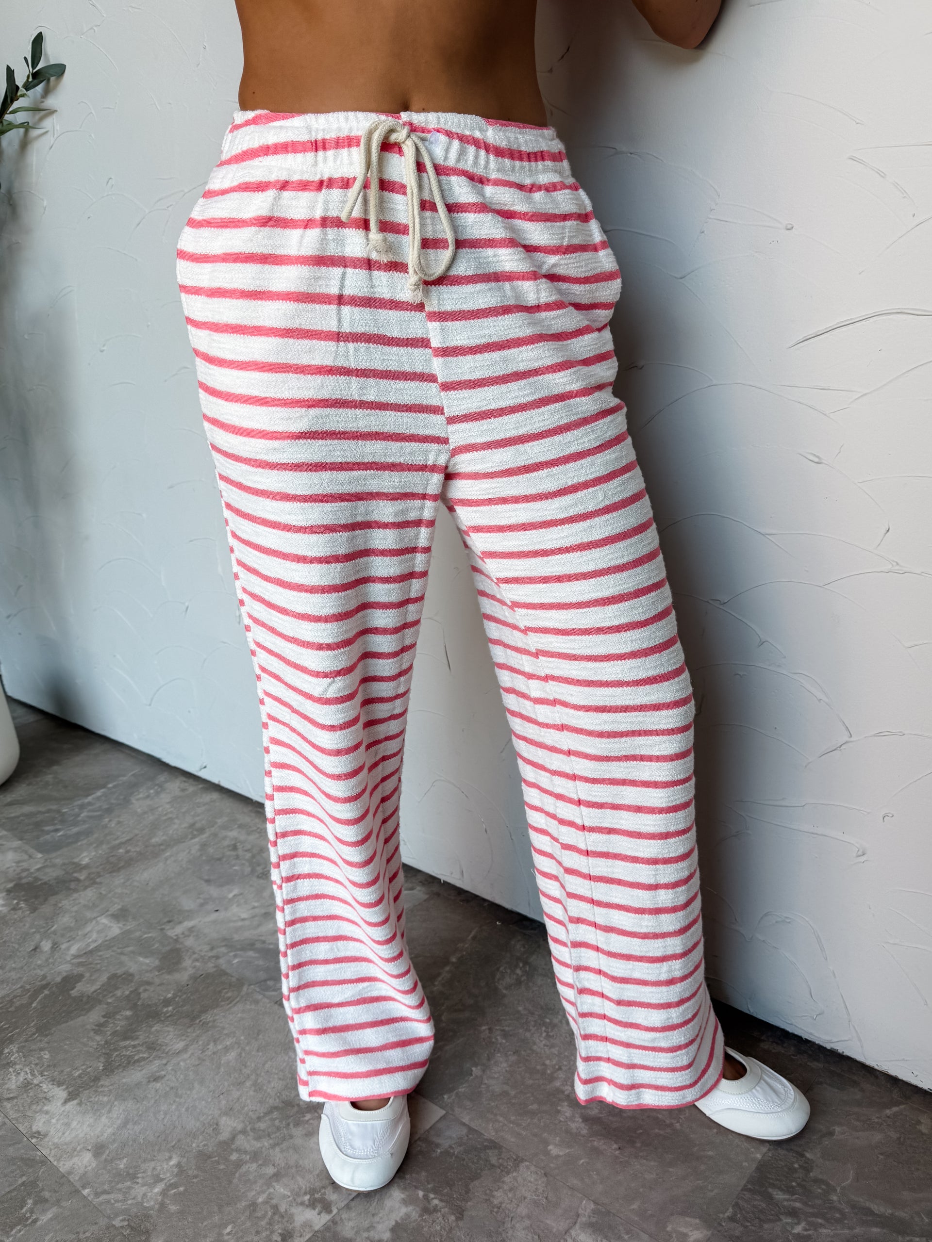 A photograph from the waist down of a model wearing high-waisted striped lounge pants against a textured white wall. The pants are made from an 87% cotton and 13% polyester blend, featuring a white textured knit base with vibrant pink horizontal stripes, an elastic waistband with a thick cream braided rope drawstring tie, and a full-length wide-leg cut, paired with white sneakers.