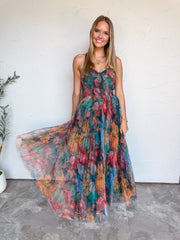A full-length photograph of a smiling young woman with straight blonde-highlighted hair swirling her dress skirt against a textured white wall. She is wearing a sleeveless maxi dress made from 100% polyester fabric, featuring a sheer pleated tulle overlay adorned with a colorful jewel-toned floral print in teal, red, pink, and orange, a smocked bustier-style bodice with thin spaghetti straps, styled with a gold heart chain necklace and delicate silver bracelets.