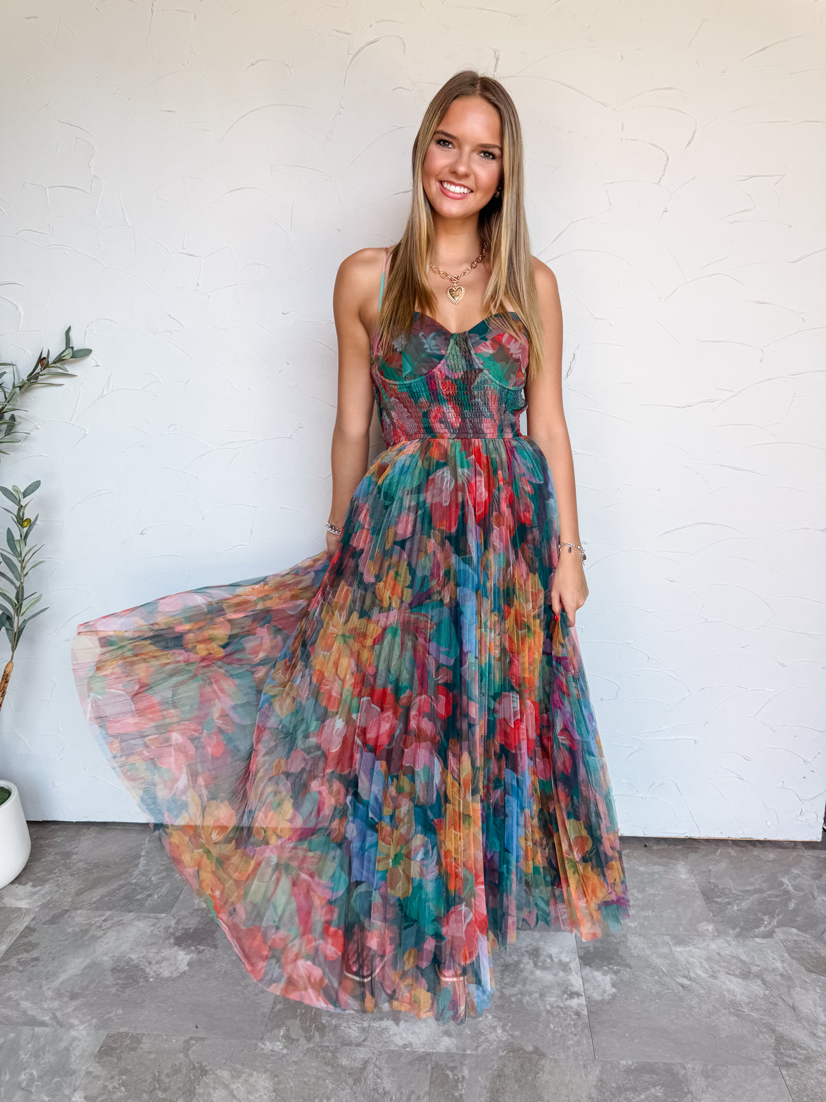 A full-length photograph of a smiling young woman with straight blonde-highlighted hair swirling her dress skirt against a textured white wall. She is wearing a sleeveless maxi dress made from 100% polyester fabric, featuring a sheer pleated tulle overlay adorned with a colorful jewel-toned floral print in teal, red, pink, and orange, a smocked bustier-style bodice with thin spaghetti straps, styled with a gold heart chain necklace and delicate silver bracelets.