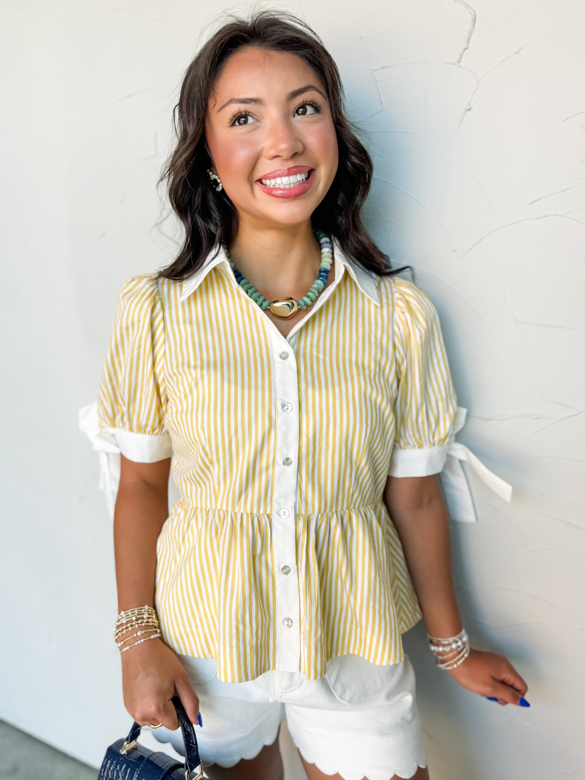 A mid-shot photograph from the chest up of a smiling brunette woman looking up and posing against a textured white wall. She is wearing a yellow and white vertically striped button-down top made from 100% cotton fabric, featuring a white collar, short puff sleeves with contrast white tie details, and a flared peplum hem, styled with white scalloped shorts.