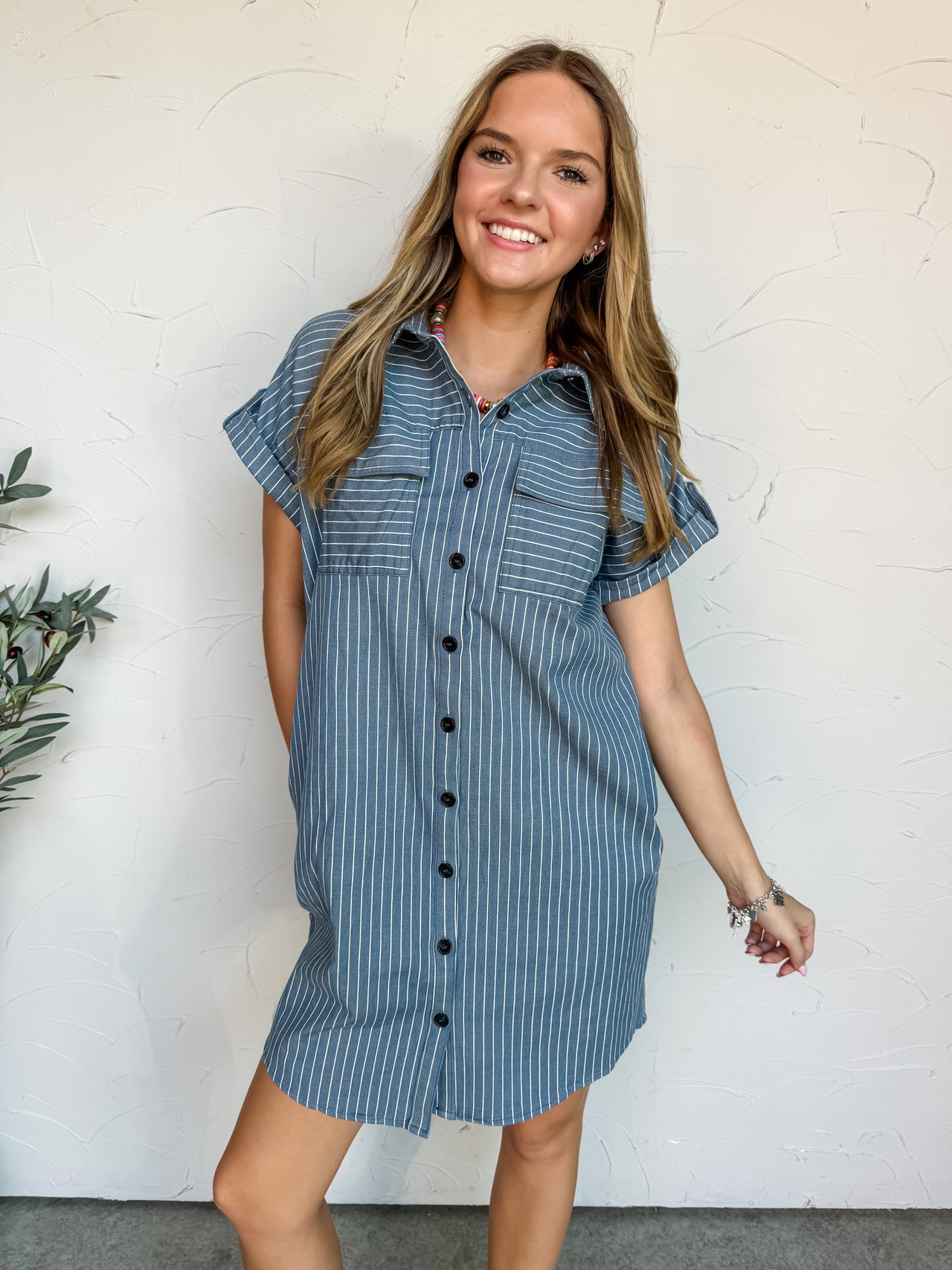 A full-length photograph of a smiling blonde woman posing against a textured white wall. She is wearing a short-sleeve button-down shirt dress made from a 65% cotton, 32% polyester, and 3% spandex blend fabric featuring a blue and white pinstripe pattern, designed with a structured collar, short cuffed sleeves, and two front buttoned chest pockets.