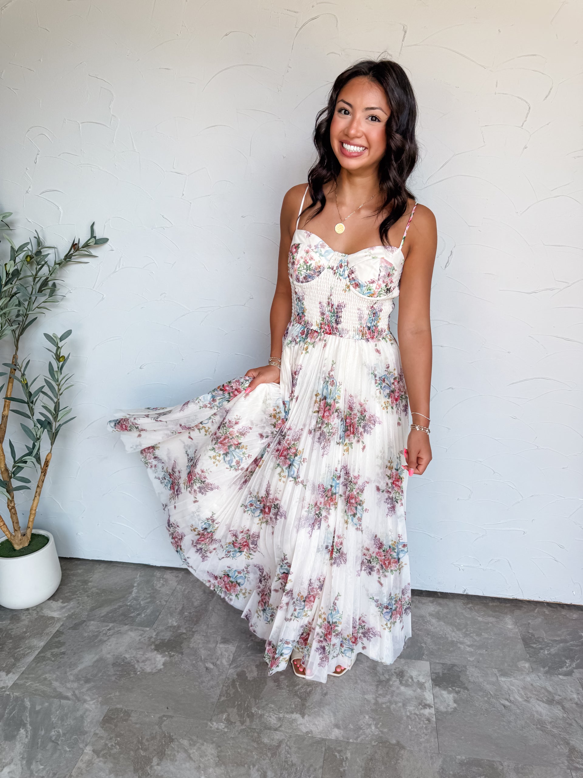 A full-length photograph of a smiling young woman with dark wavy hair fanning out her dress skirt against a textured white wall. She is wearing a sleeveless maxi dress made from 100% polyester fabric, featuring a sheer pleated tulle overlay with an ivory background and a delicate floral print in pink, blue, and green, a smocked bustier-style bodice with thin spaghetti straps, styled with a gold pendant necklace and gold beaded bracelets.